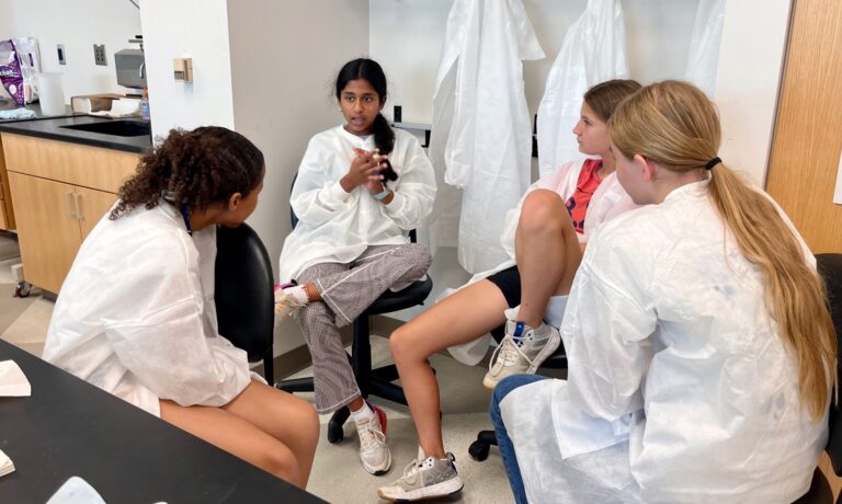 four girls, wearing lab coats, sitting in a circle in a laboratory