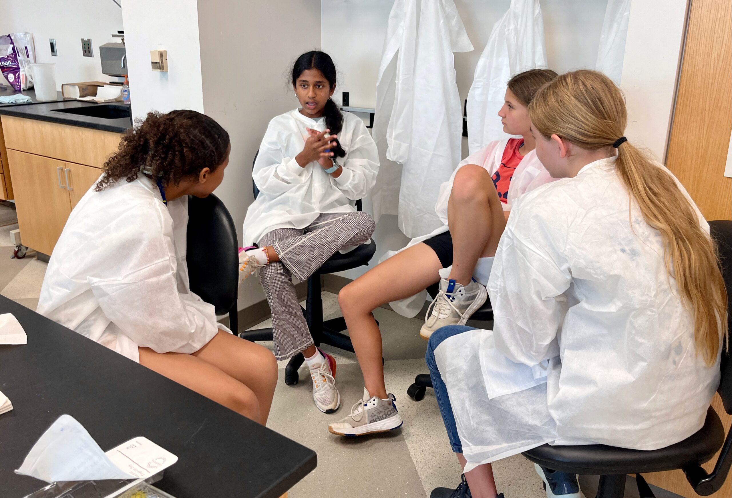 four girls, wearing lab coats, sitting in a circle in a laboratory