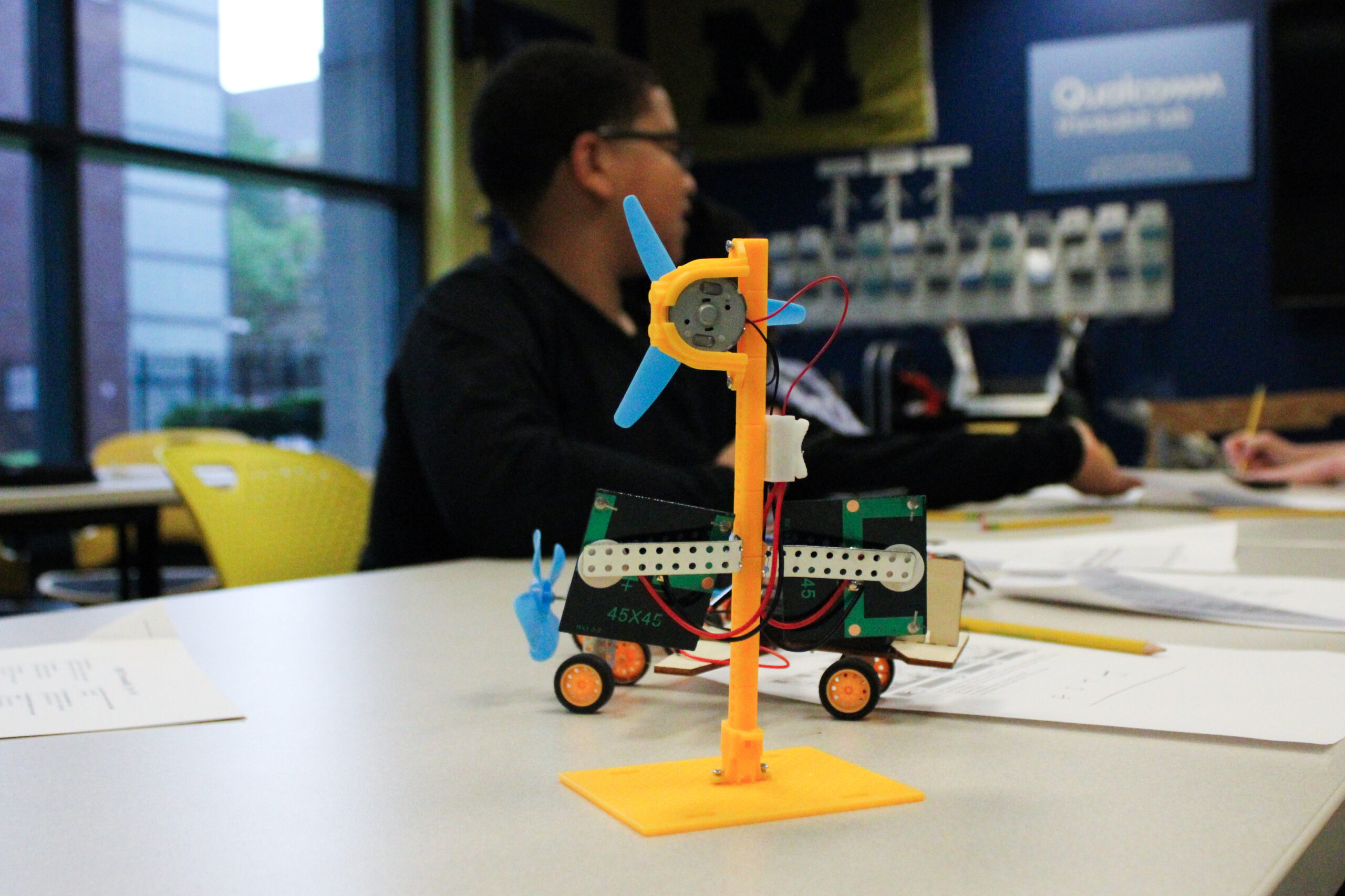 A student's model wind turbine, built with a blue propeller on an orange 3D-printed stand, displayed on a classroom table during a WISE program in Detroit.