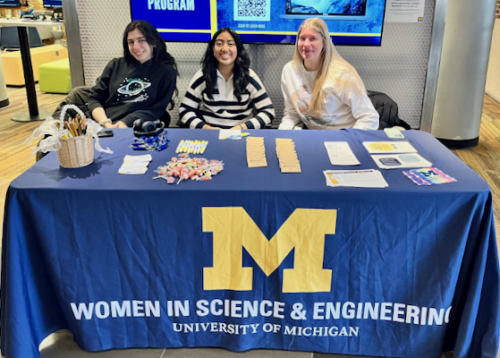 Three WISE student volunteers sit behind an information table with a Women in Science & Engineering tablecloth.
