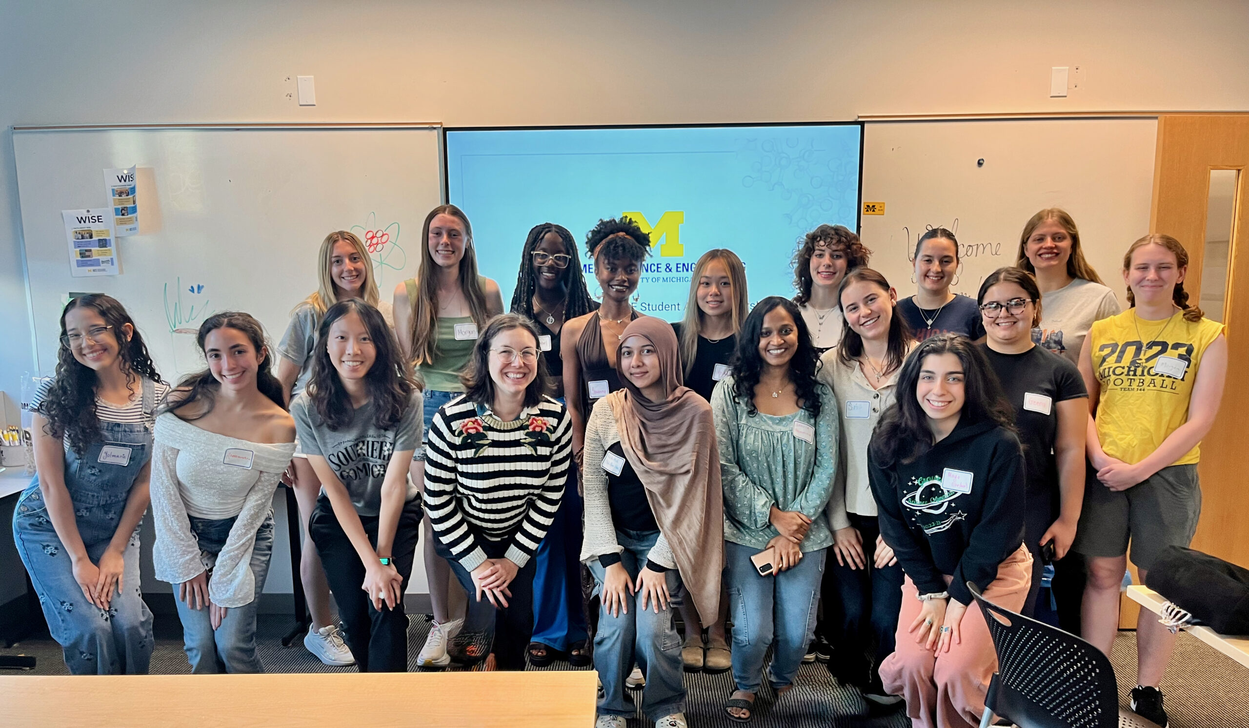 A large group of WISE students and staff pose in a classroom in front of a screen displaying a Women in Science & Engineering presentation.