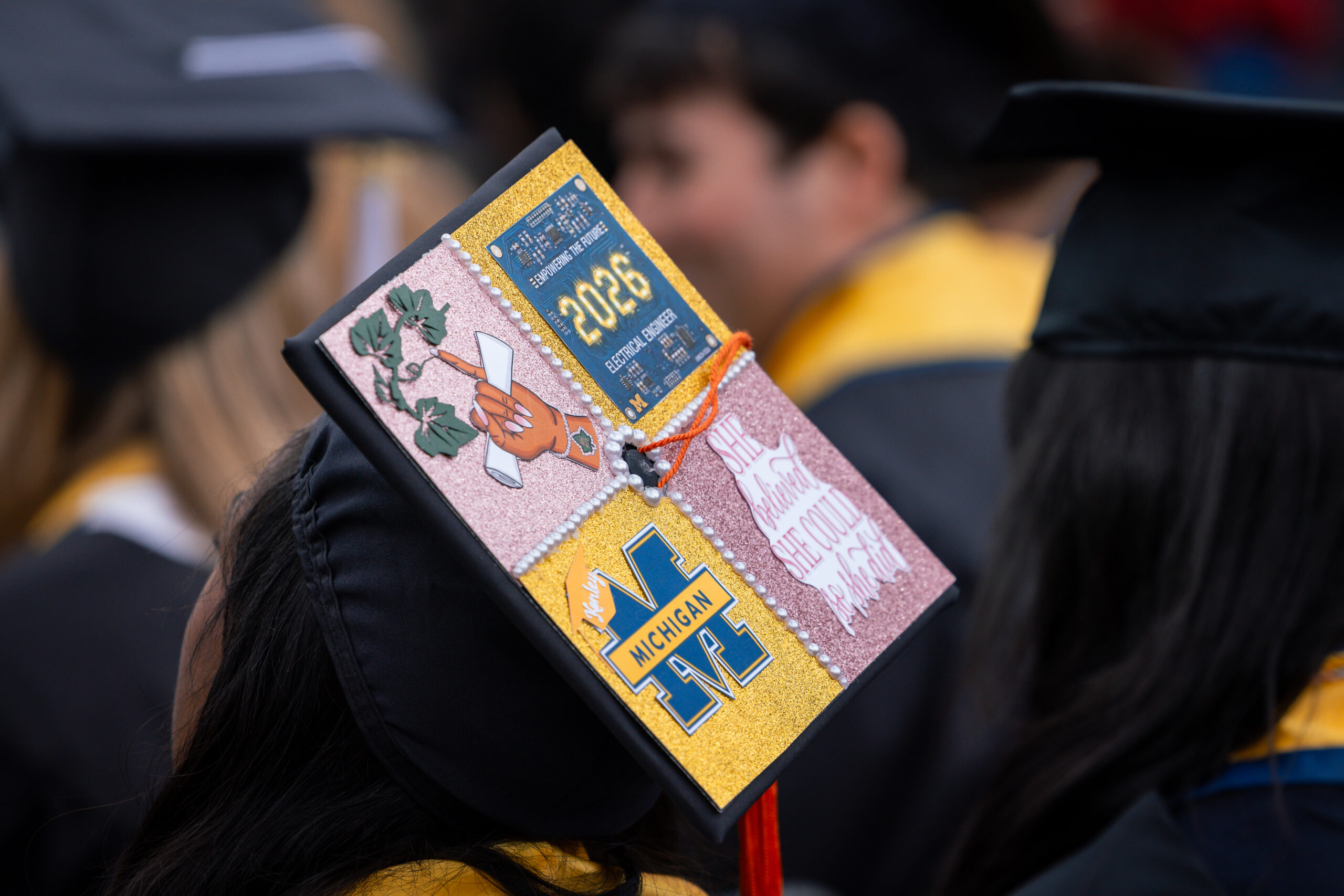 Close-up of a decorated graduation cap reading "2026 Electrical Engineer," decorated with a circuit-board design, a Michigan block M, and the phrase "She believed she could so she did," worn at commencement.