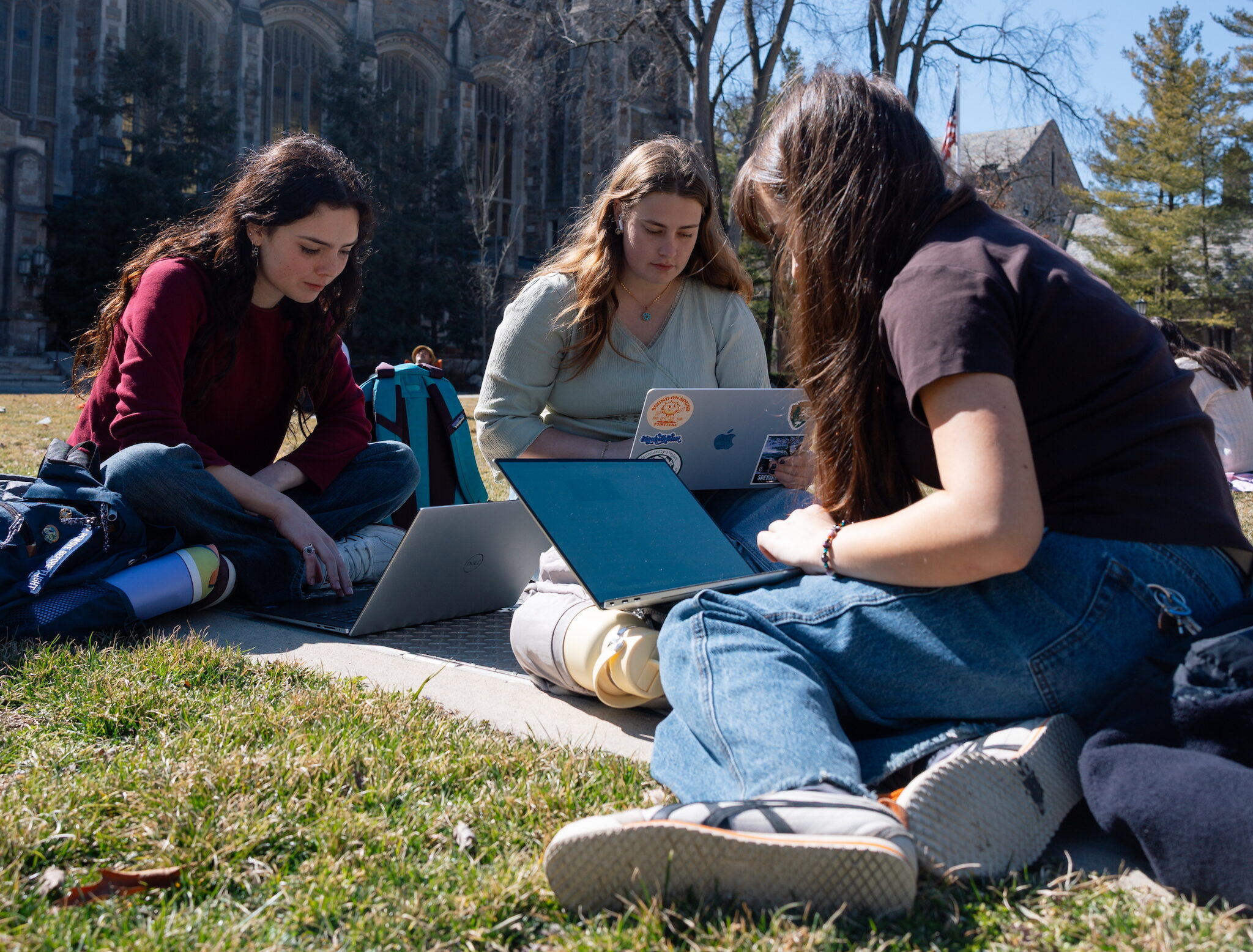 Three female college students sit facing each other on the grass outside the law school studying, using laptops.