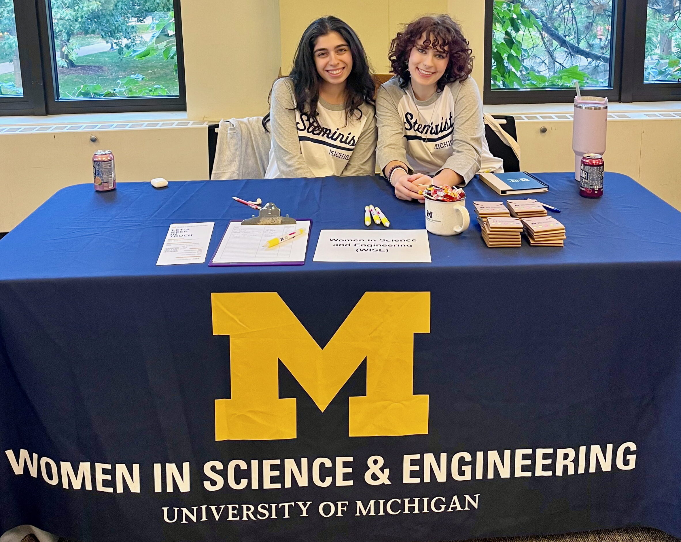 Two female students, wearing shirts with the word Steminist across the chest sit behind a table with a blue table cloth. The table cloth has a Block M and the text on the table cloth says Women in Science and Engineering, University of Michigan