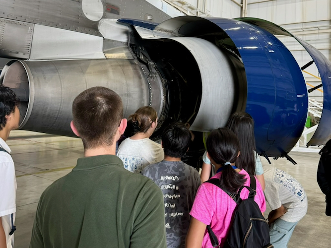 A group of middle school campers and two camp instructors look at a jet engine being repaired in a hangar at Detroit Metro Airport.