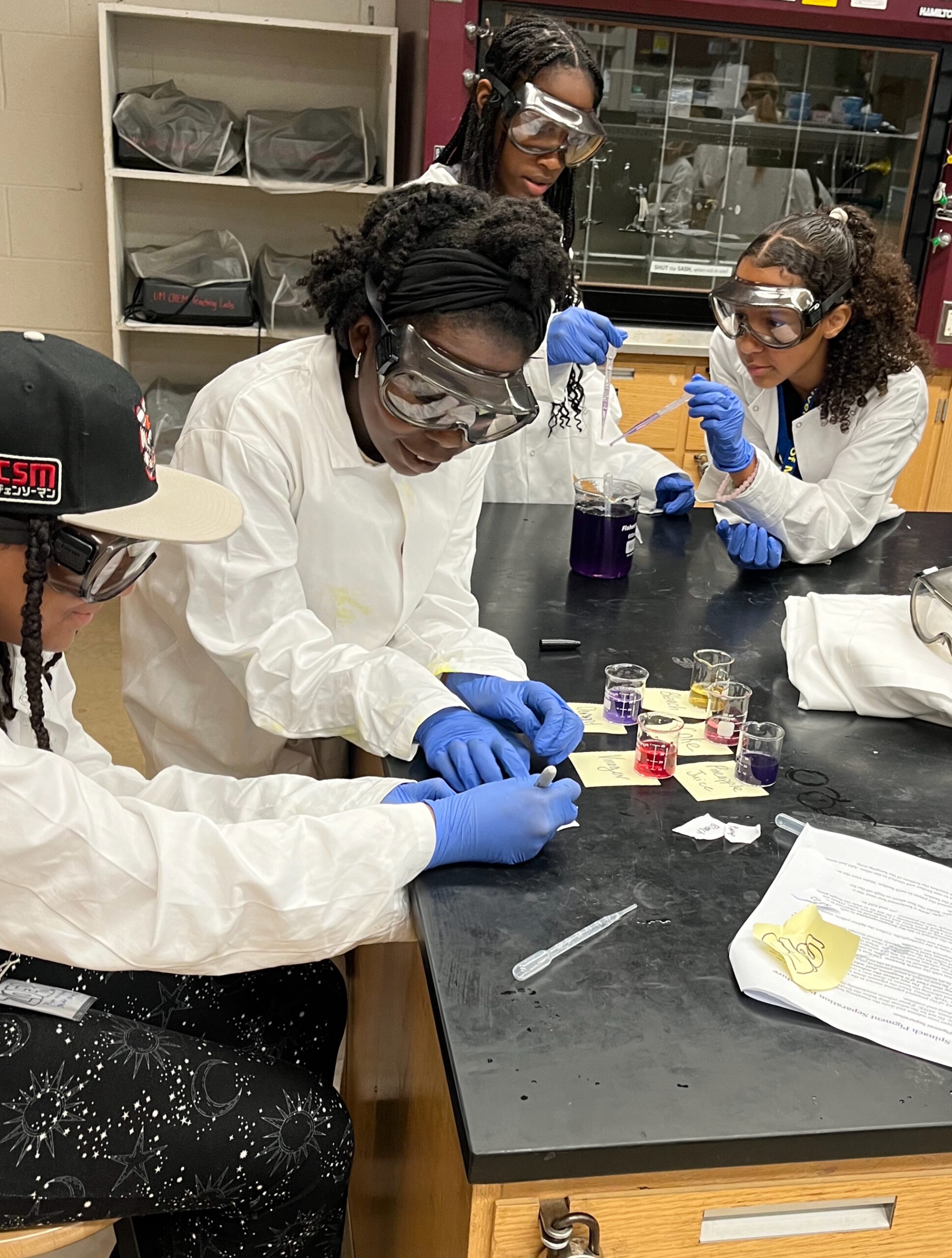 Four middle school scientists wearing lab coats, goggles and gloves conduct experiments at WISE's summer camp.